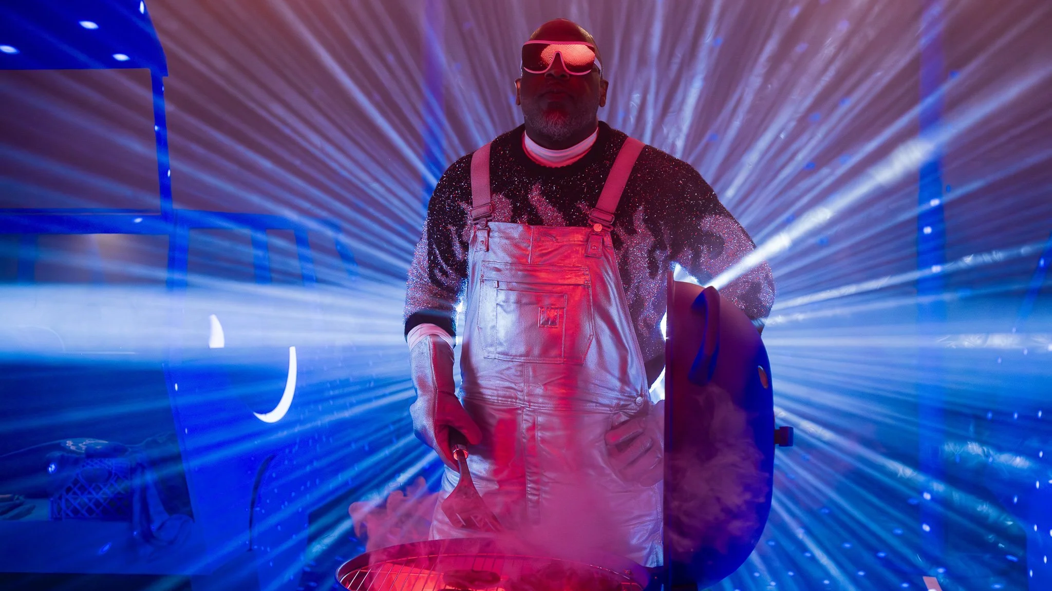 A person wearing reflective sunglasses, glittery shirt, and overalls holding a tool, surrounded by colorful laser lights and smoke.