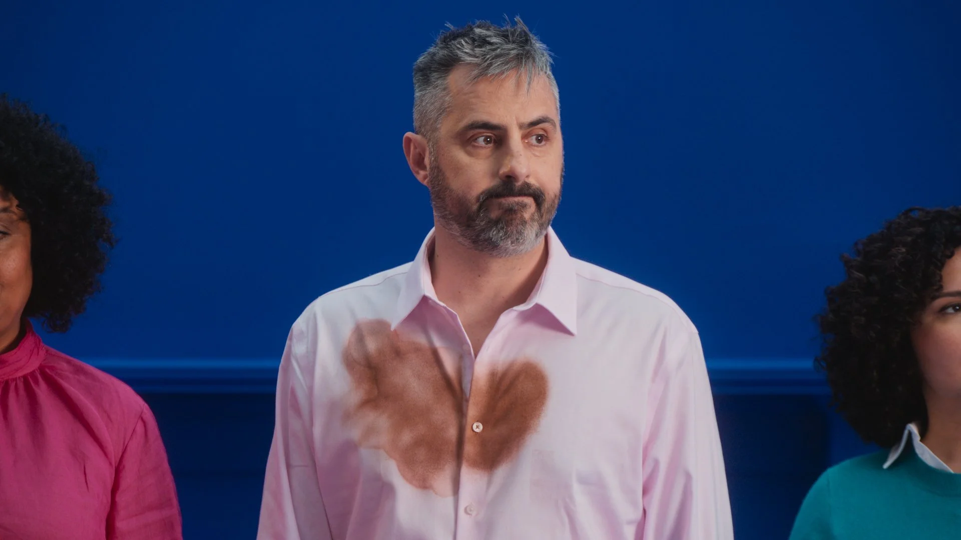 A man with gray hair and a beard standing between two women, wearing a white shirt with a brown stain on it, against a blue background.