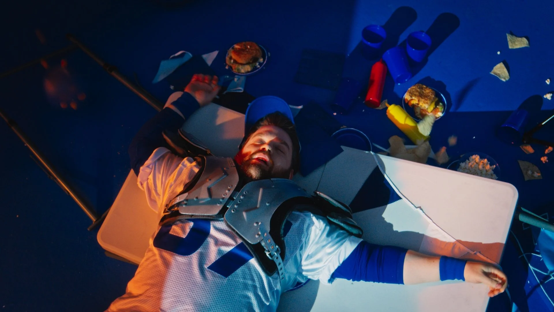 A man with a beard lies on the ground dressed as an astronaut with snacks and cups scattered around him on the floor, under blue and red lighting.