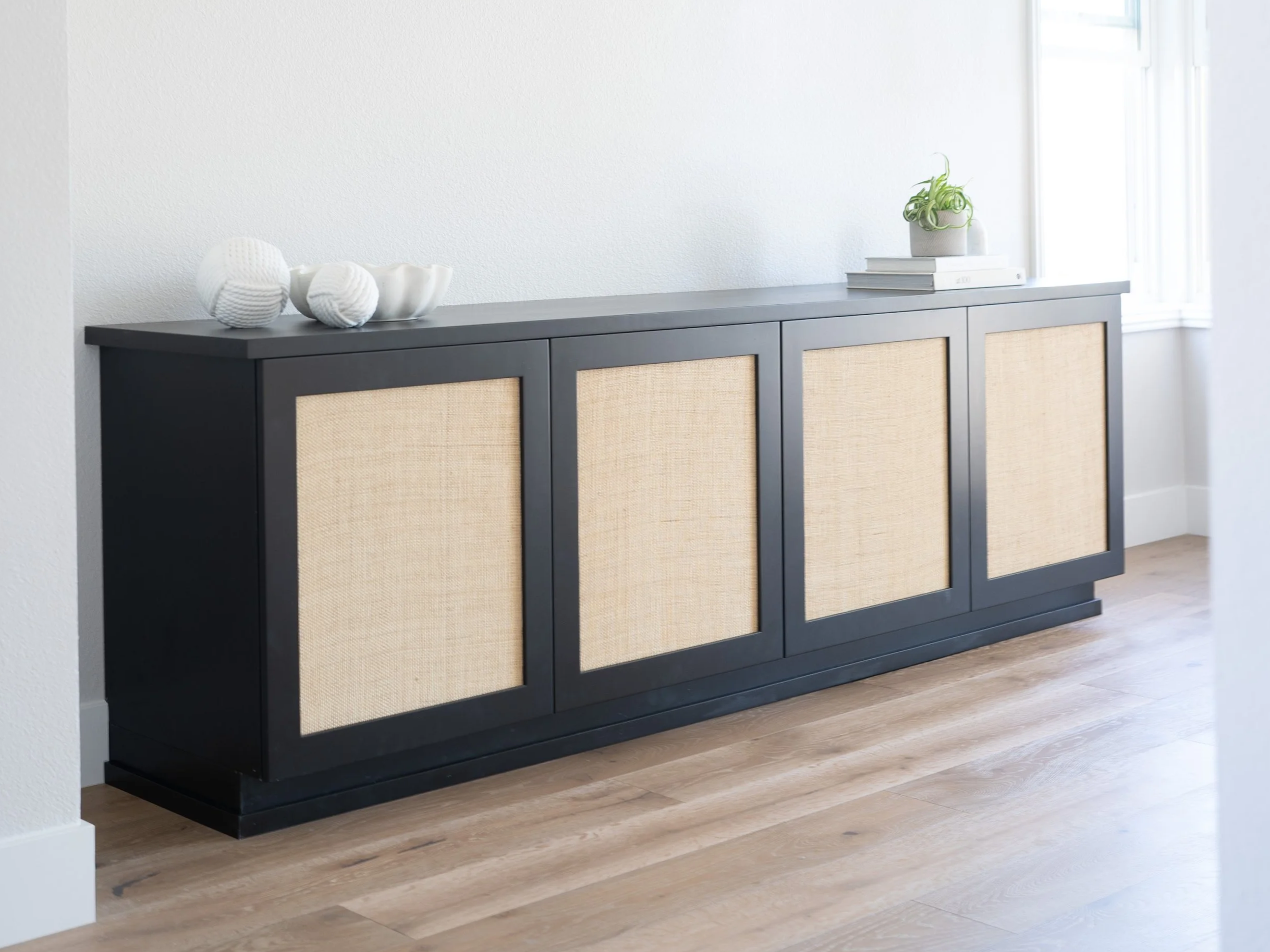A custom designed and constructed built in entertainment console with inset woven raffia cabinet doors is placed against a soft gray wall near a window. On top are stylish decorative ceramic vases, a small potted plant, and a stack of design books.