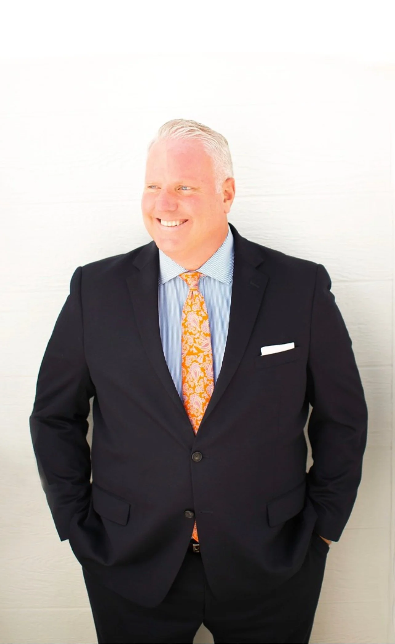 Man wearing a black suit, light blue shirt, orange patterned tie, and white pocket square, smiling and standing against a plain white background.