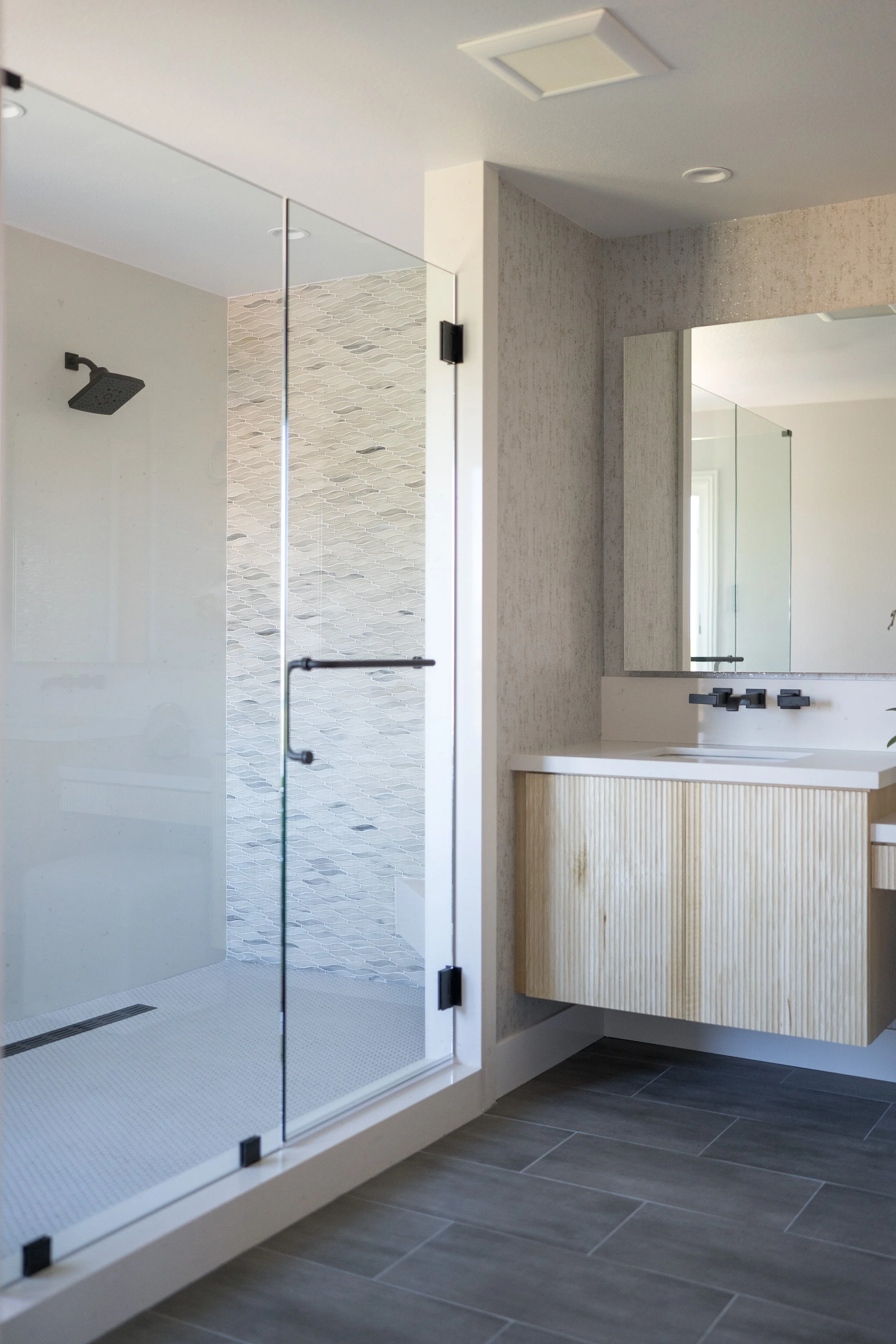 Modern and spacious primary bathroom with oversized walk-in shower featuring solid quartz slab walls, dual shower heads with a handheld showerhead, wall controls, and a custom white oak fluted vanity, quartz counters, and designer wallpaper.