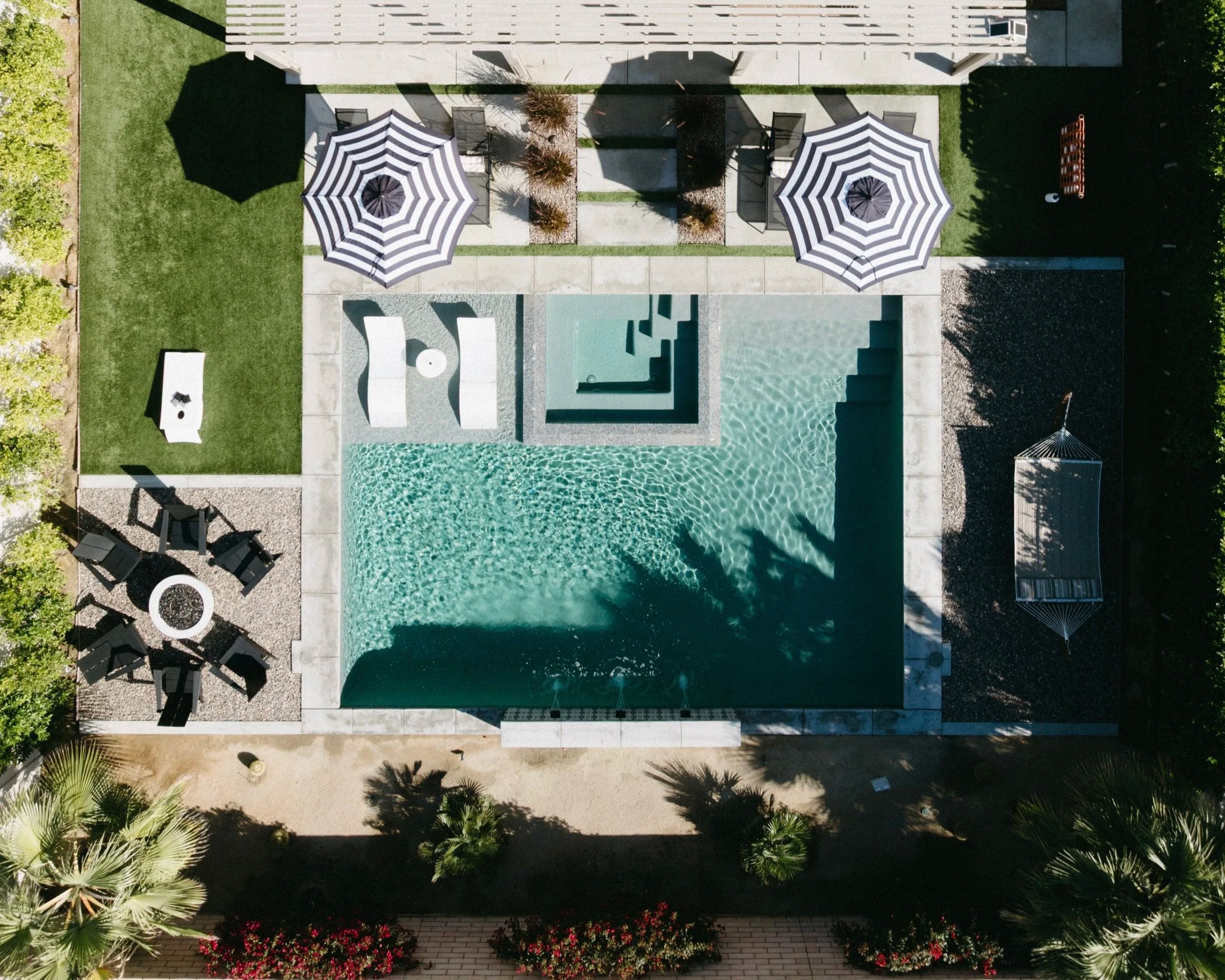 Top-down view of a backyard with a swimming pool, two striped umbrellas, lounge chairs, and surrounding lush greenery.