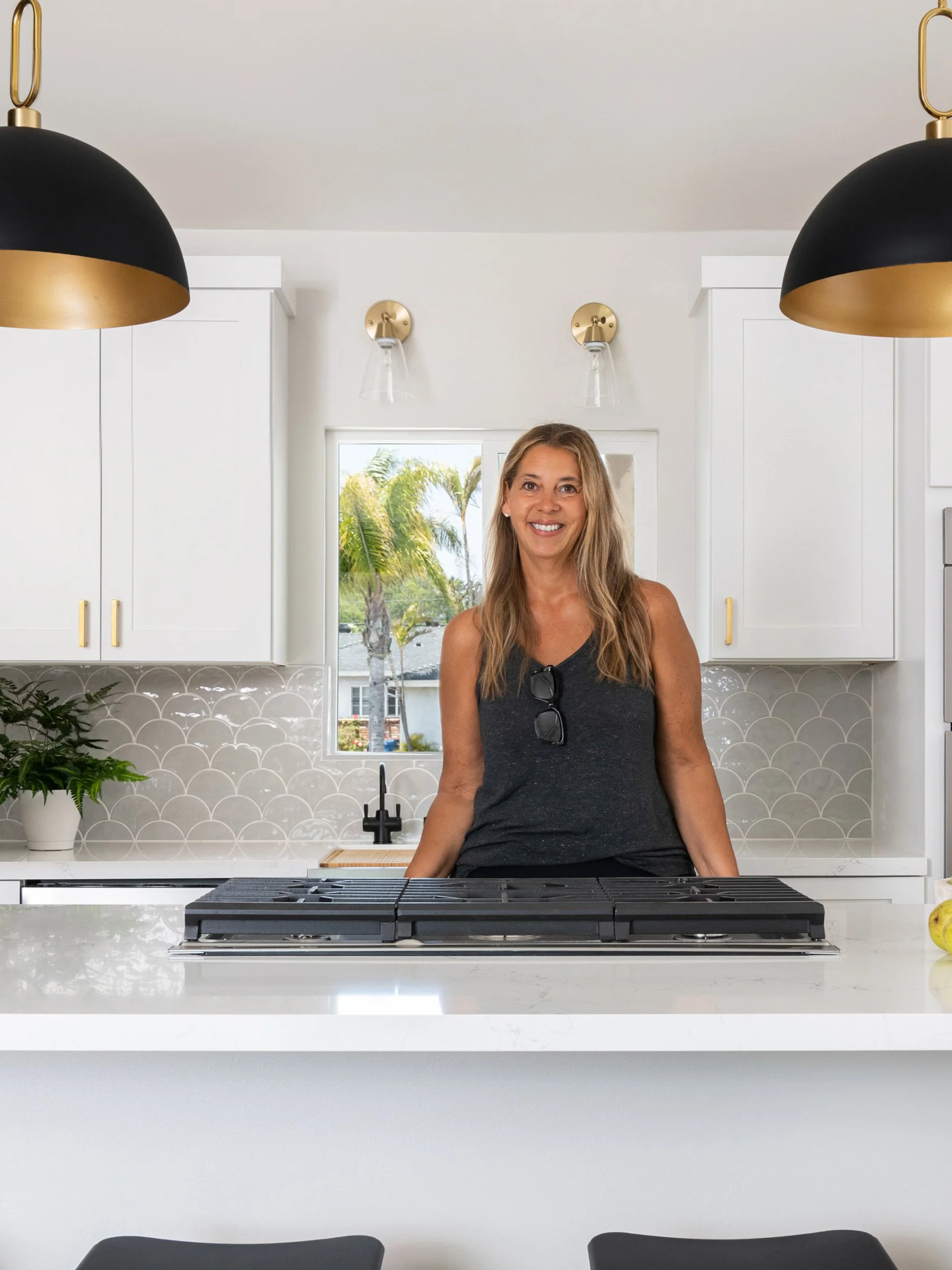 A woman with long blonde hair, wearing a dark sleeveless top and sunglasses hanging on her shirt, standing in a modern kitchen with white cabinets, black and gold hanging lamps, a window with a view of a palm tree outside, and a gas stove in the foreground.