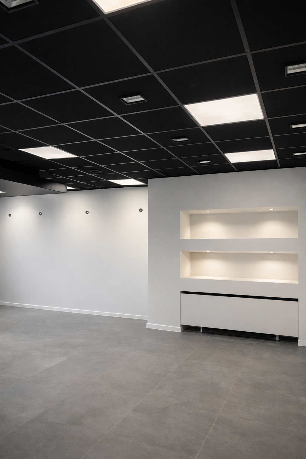 Empty modern interior with black ceiling tiles and recessed lighting, white walls with built-in shelves, and gray tiled flooring.