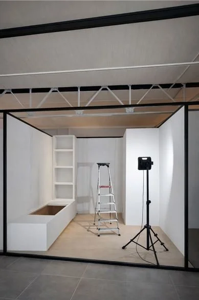 Empty photo studio with white walls, a small ladder, a shelving unit, a rectangular bench, and a light setup on a stand.