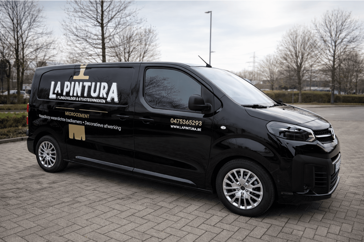 Black van with branding for 'La Pintura' including contact number and website, parked on a paved lot with trees and overcast sky in background.
