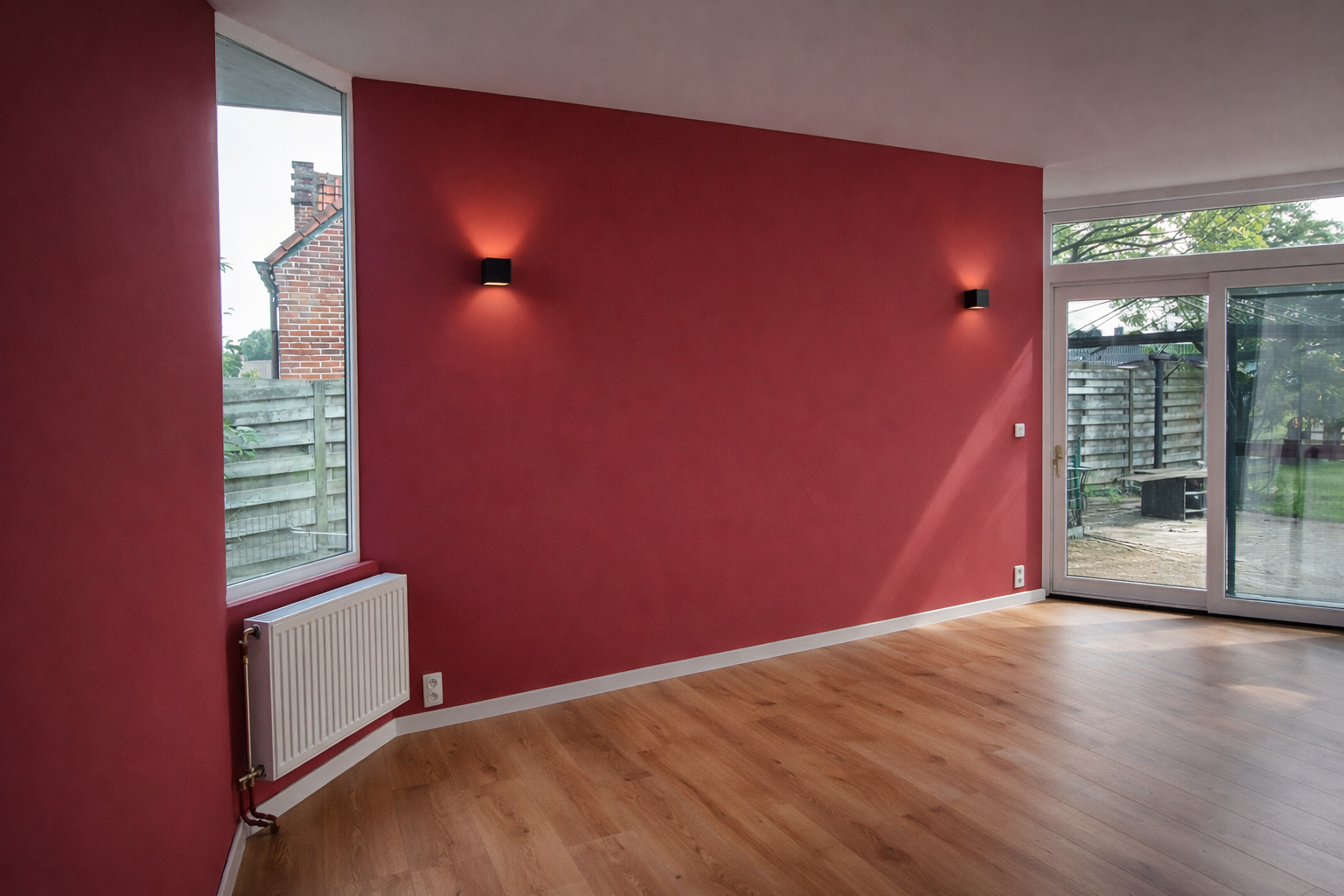 Lege woonkamer met een rode accentmuur, houten vloer, groot raam en een glazen deur die naar de achtertuin leidt, met een buitenpatio zichtbaar.