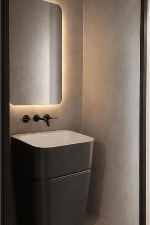 Modern bathroom with a white pedestal sink, black fixtures, and a backlit mirror on a gray wall.
