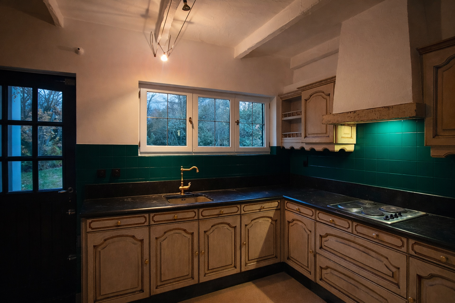 Keuken met houten kasten, een groene betegelde achterwand, een zwart werkblad, een messing kraan, kookplaat, een raam met uitzicht op bomen buiten en een deur links met meerdere glazen ruitjes.