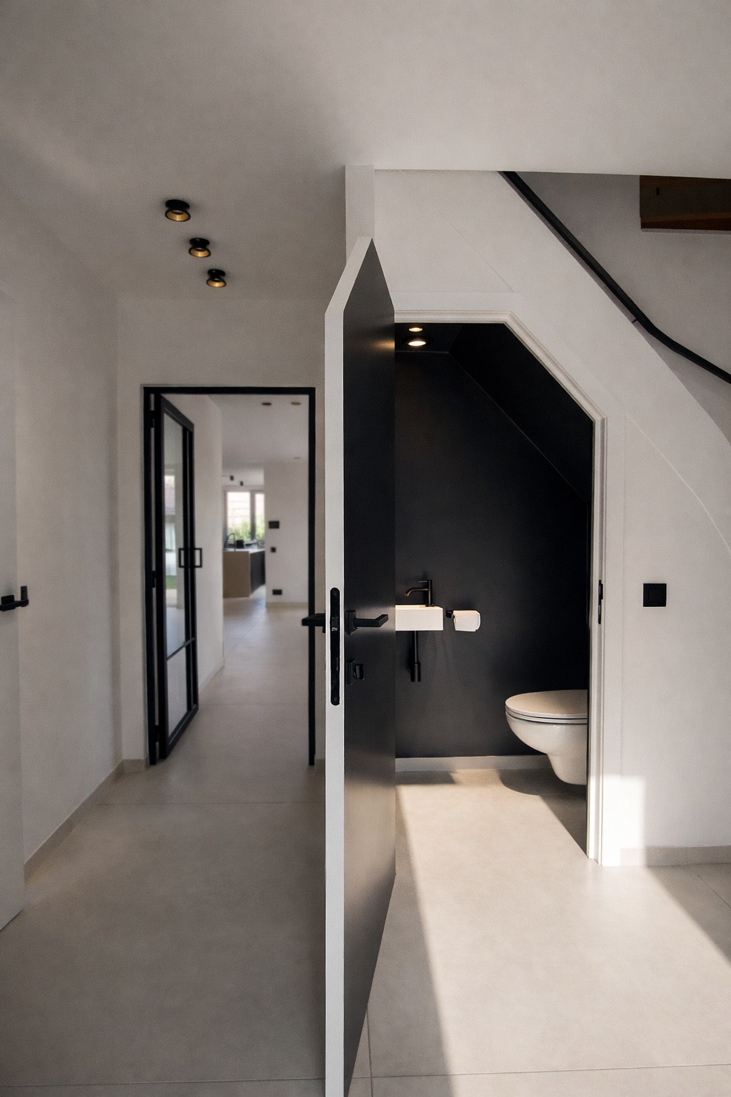 Modern interior view showing an open black door leading to a small bathroom with black walls, and a white toilet and sink. The background features a hallway with black-framed glass doors and a living area with large windows.