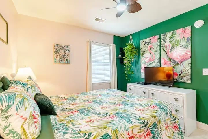Tropical-themed bedroom in a Myrtle Beach SC rental condo at Waterway Village with floral bedding, green accent wall, flamingo wall art, and ceiling fan.