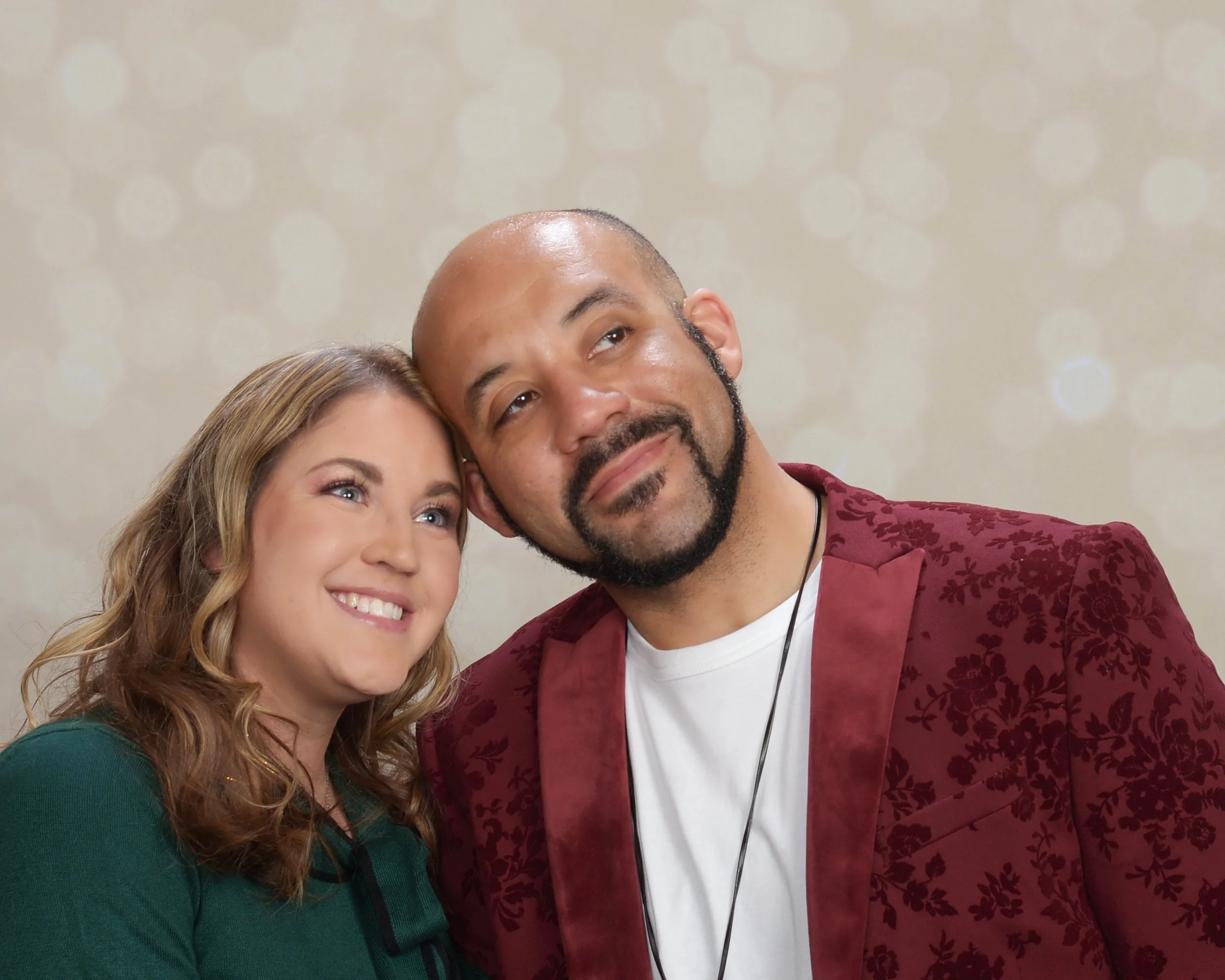 Elizabeth Muckensturm and Roca Williams smiling with heads together, portrait against a soft beige background with light bokeh details.