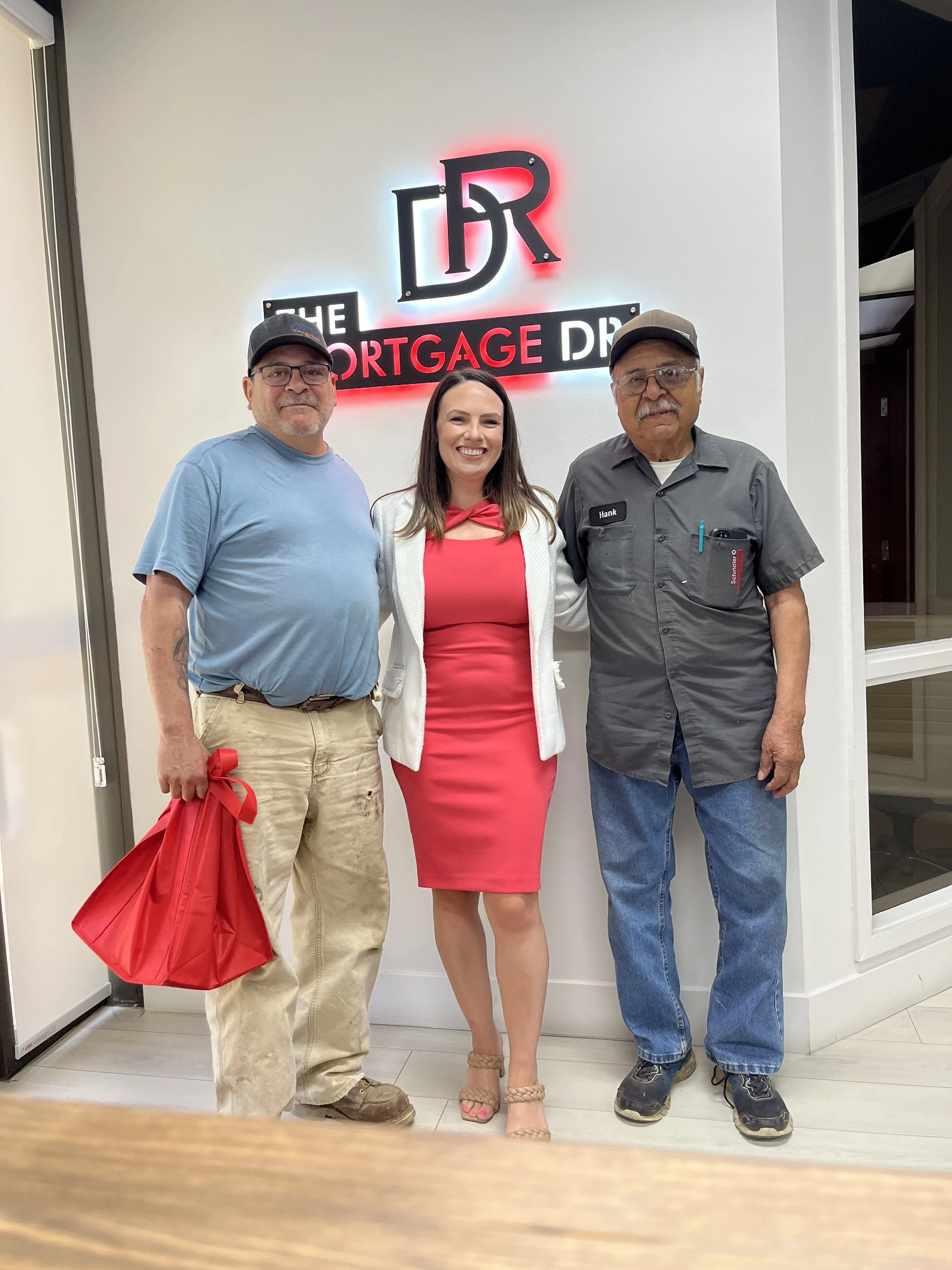Three people standing inside a building in front of a sign that reads "The Mortgage Dude." The person on the left is a man with glasses, wearing a blue t-shirt, khaki pants, and holding a red bag. The woman in the middle is wearing a red dress, a white blazer, and nude high heels. The person on the right is a man with glasses, wearing a gray shirt with a name tag that says "Hank," and blue jeans.