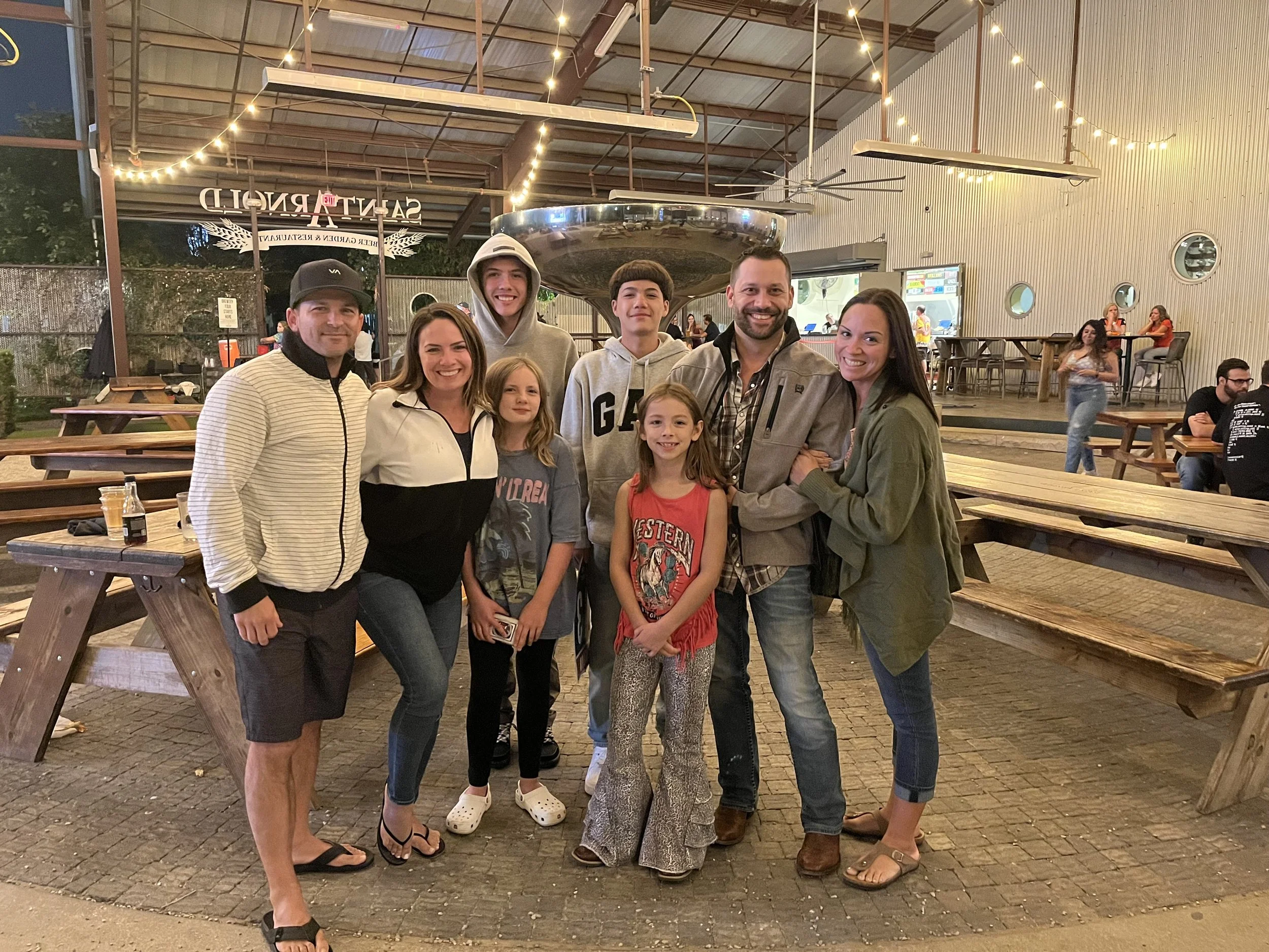 A group of nine people, including children and adults, smiling and posing together inside and outside a restaurant or bar with string lights overhead and picnic tables.