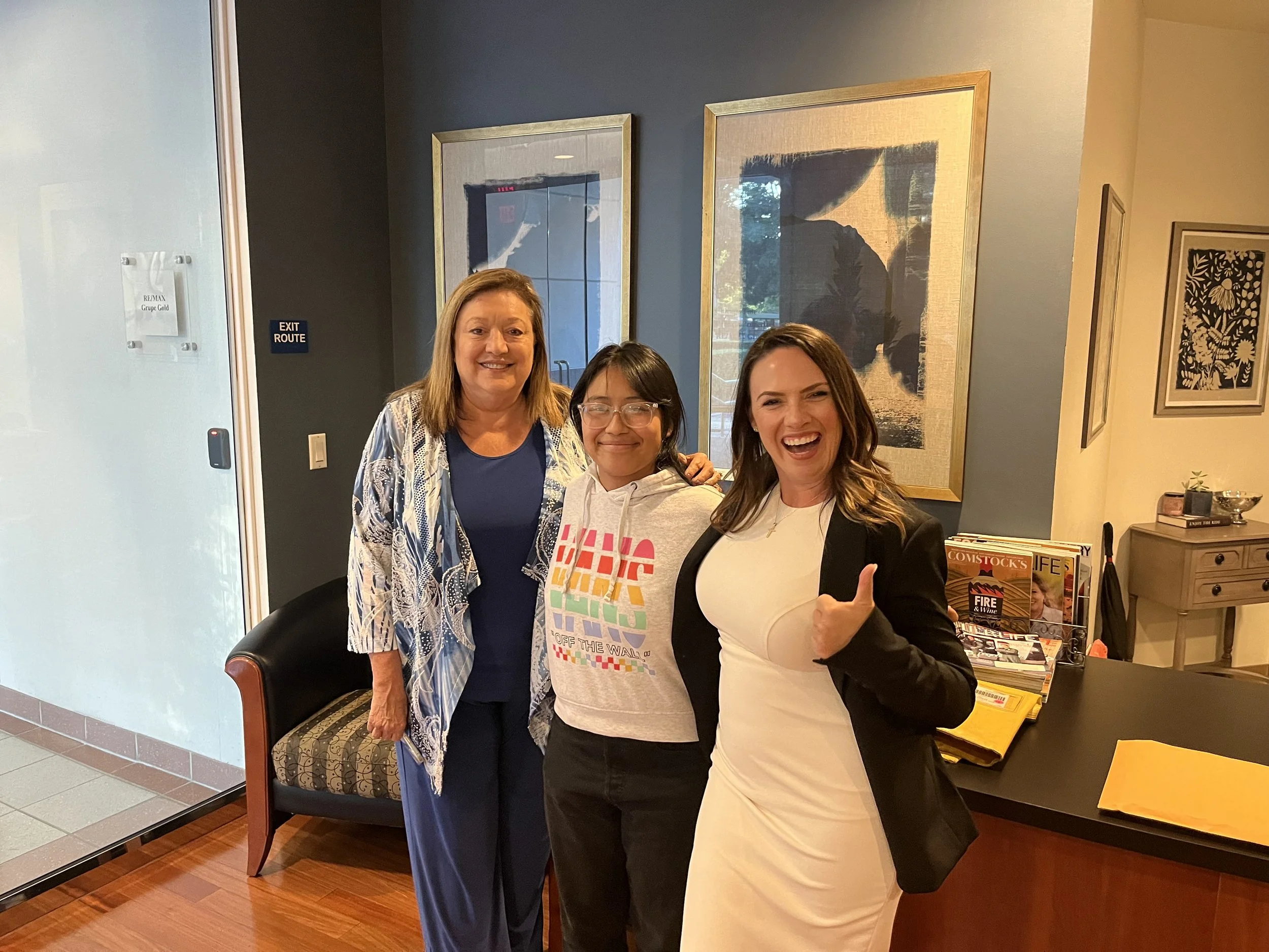 Three women standing inside a lobby area, smiling. The woman on the left is wearing a patterned blue and white kimono over a blue top. The woman in the middle is wearing glasses and a colorful hoodie. The woman on the right is wearing a white dress and a black blazer, giving a thumbs up.