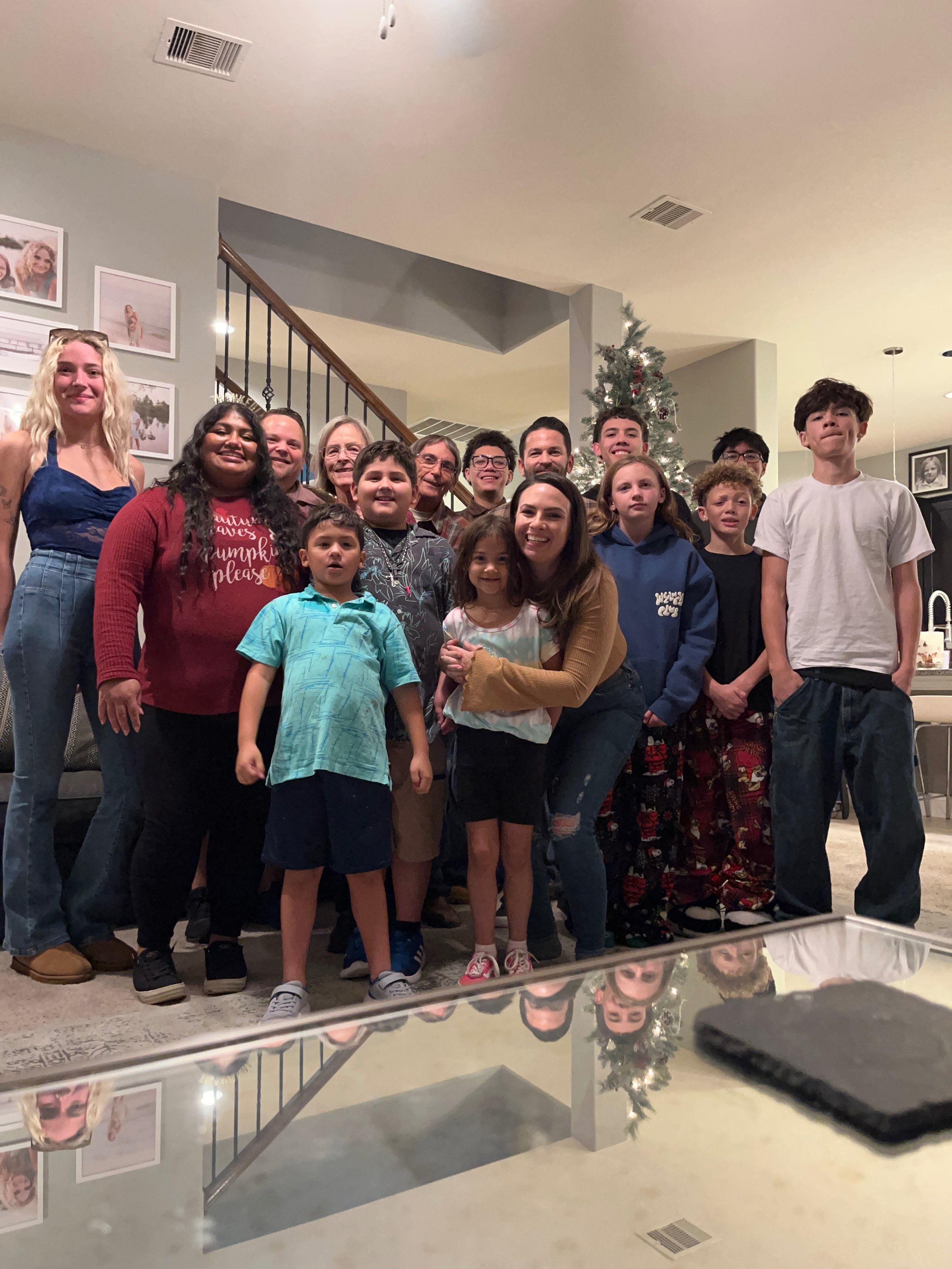 A group of people, including children and adults, gathered together for a photo in a living room with a Christmas tree in the background.