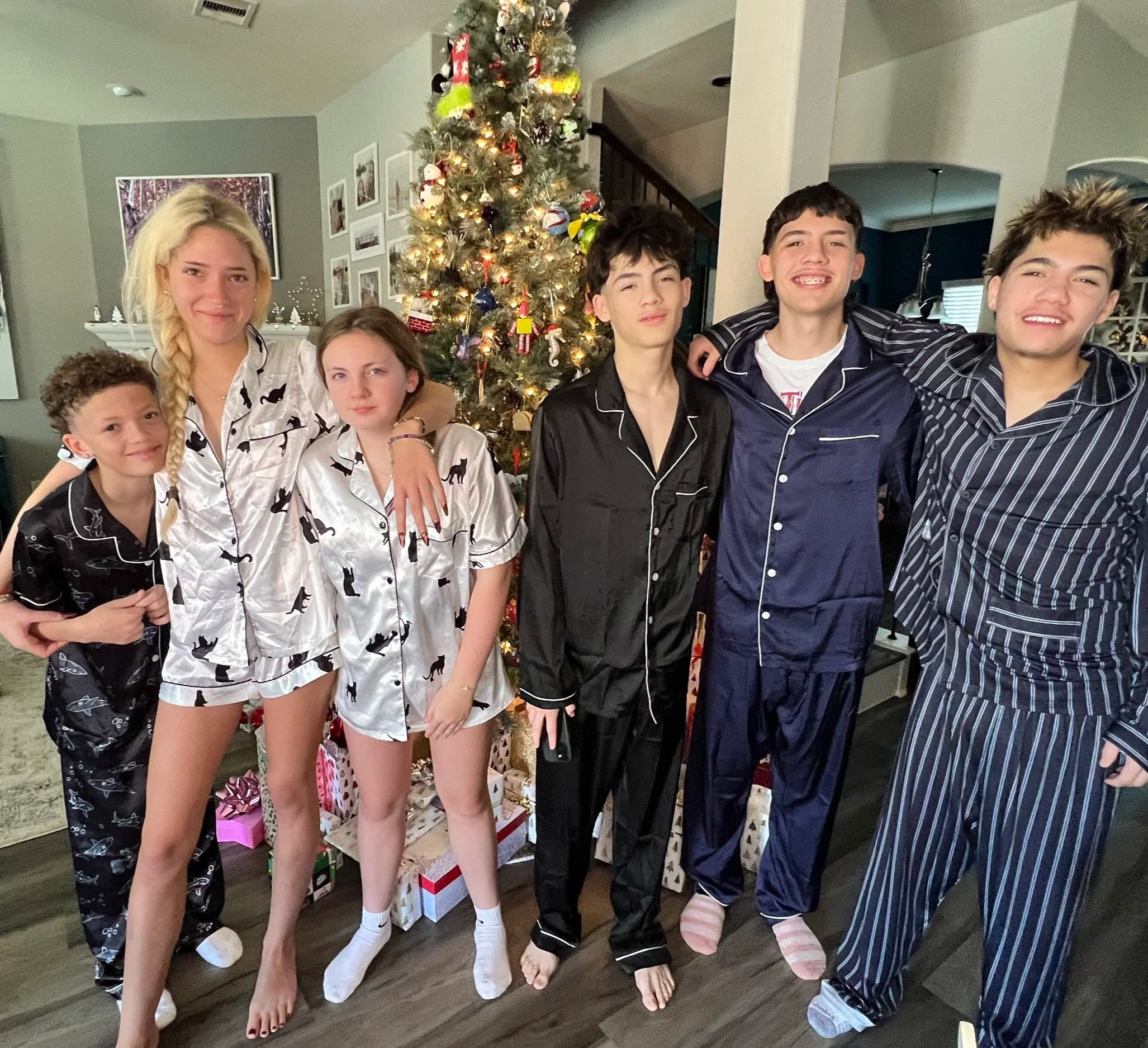 Group of six kids and teens in pajamas standing in front of a decorated Christmas tree with gifts underneath.
