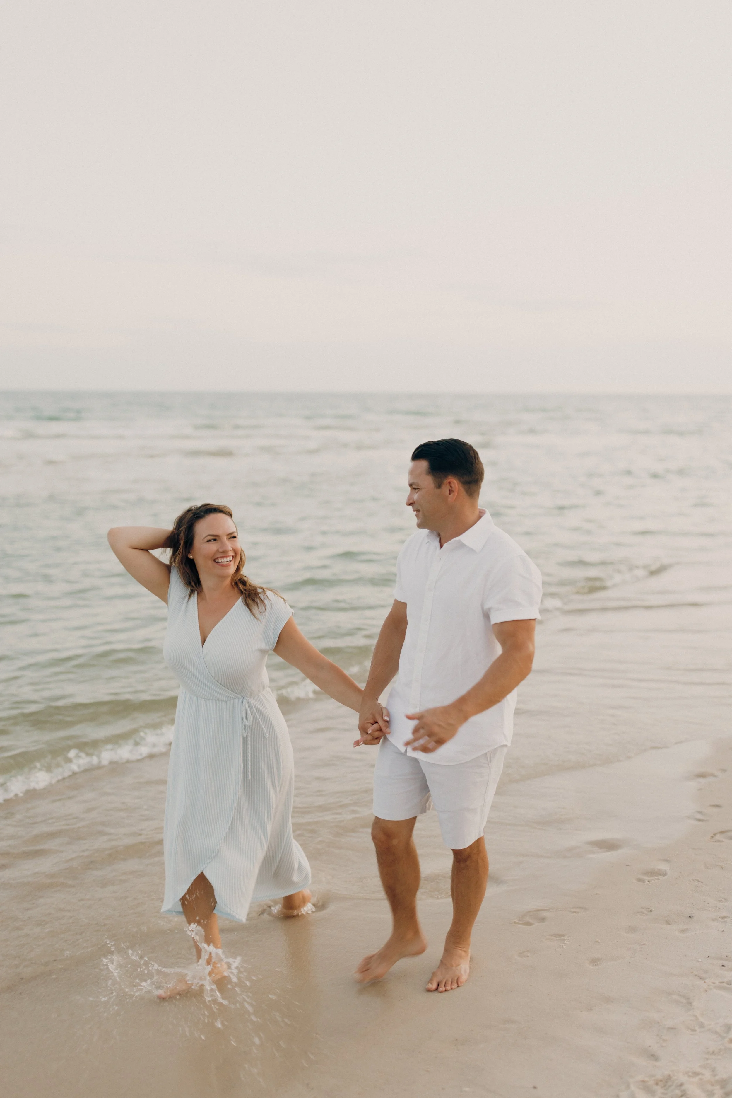 A smiling woman and man holding hands and walking along the beach, wading in the water, and enjoying each other's company.