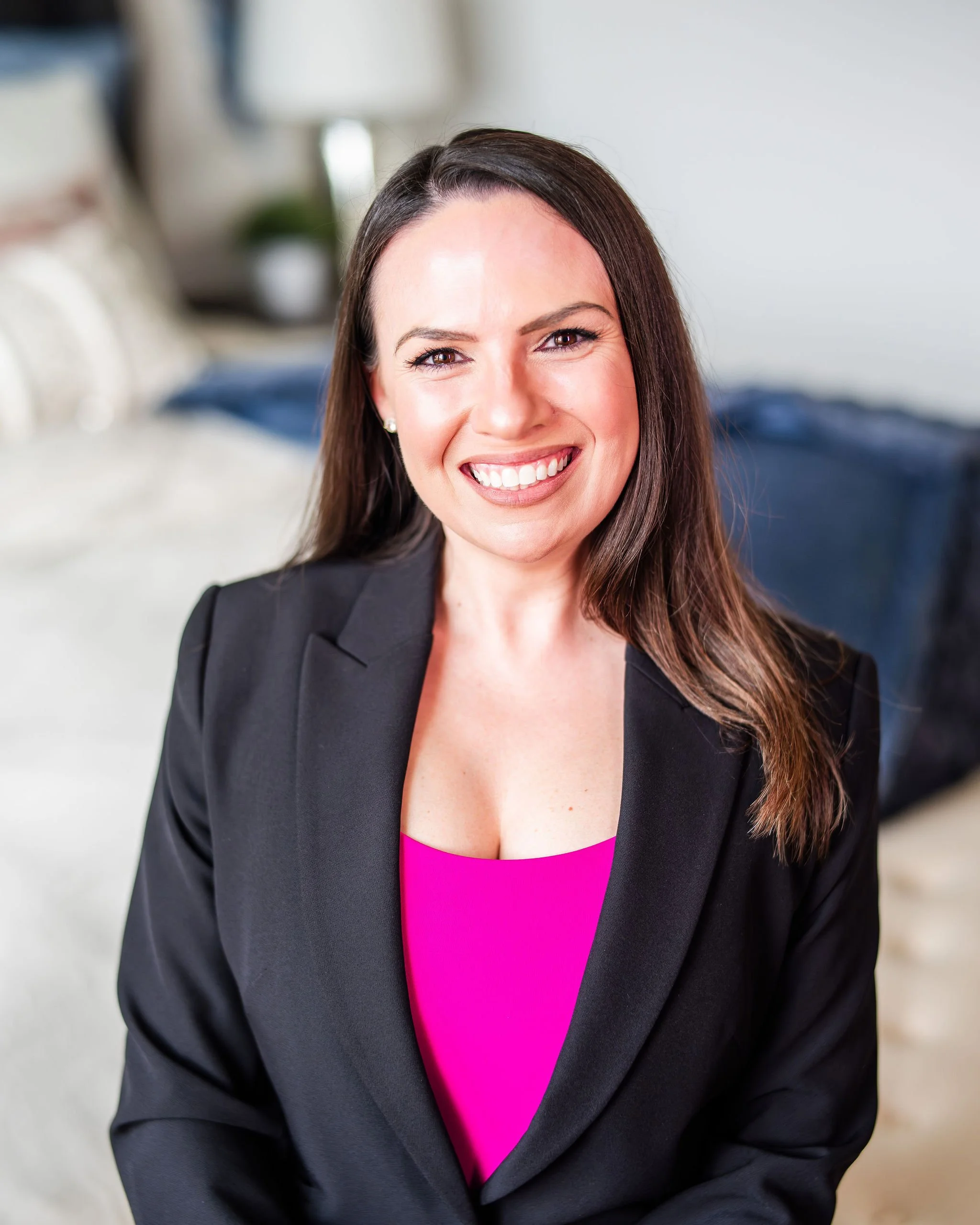 Angela Jimenez smiling with long dark hair wearing a black blazer and a bright pink top, sitting in a cozy living room.