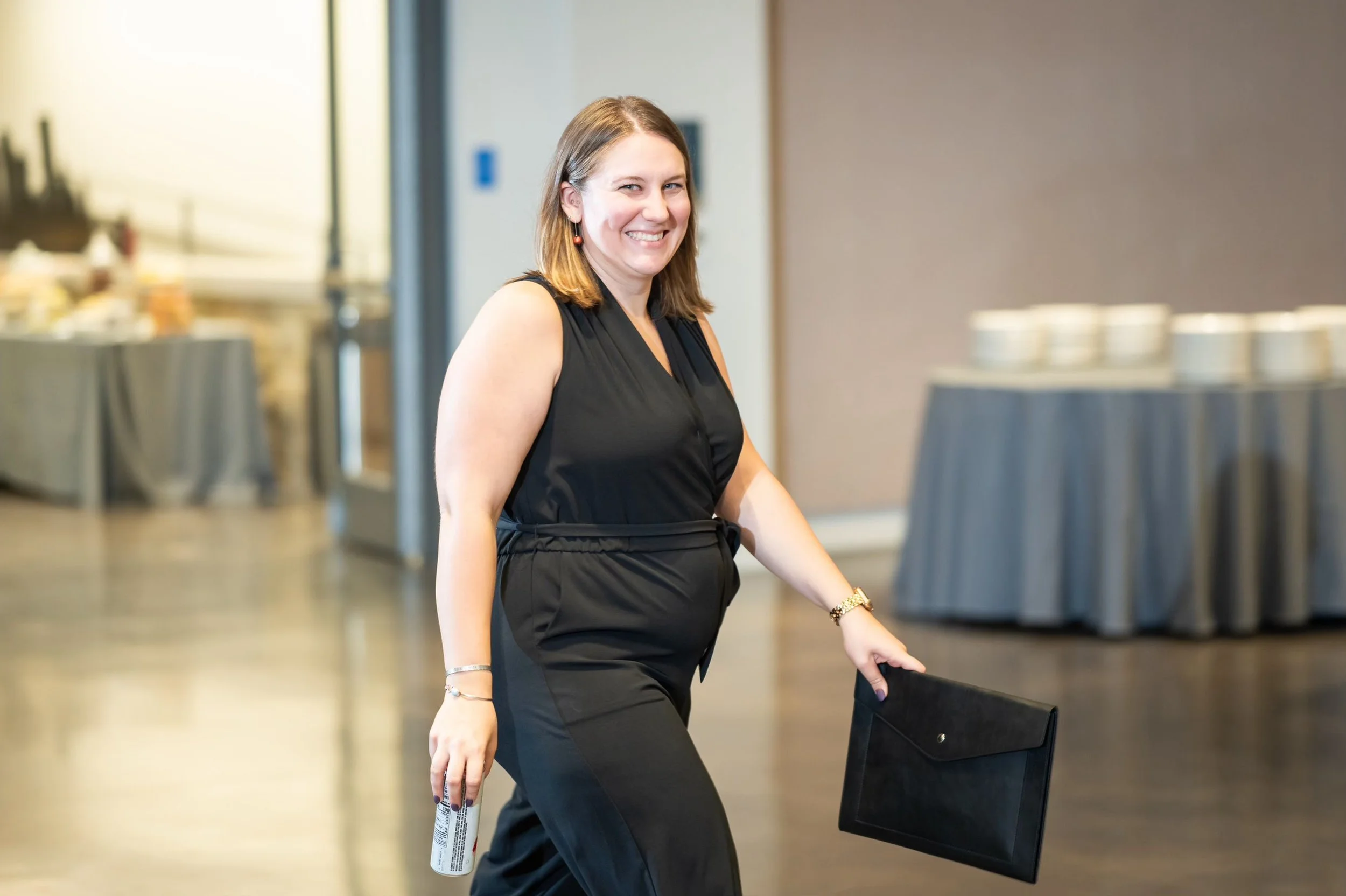 Event planner with black jumpsuite and black portfolio smiles at wedding photographer on her way to deliver a beverage to the bride