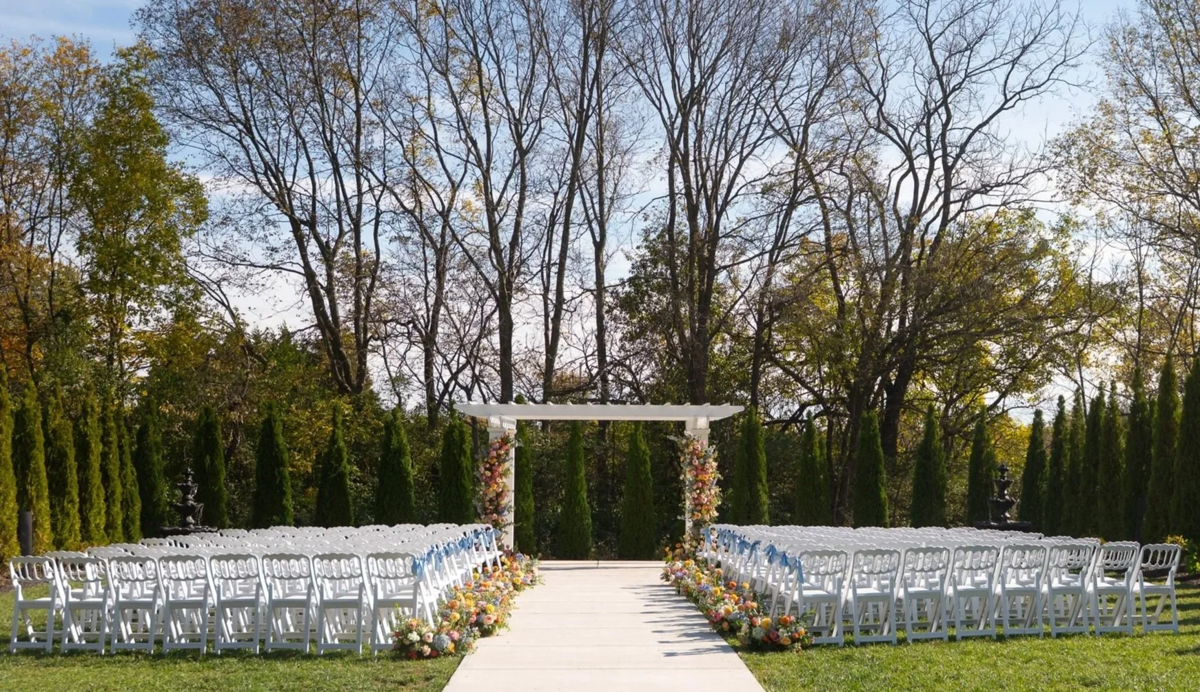 Colorful wedding ceremony featuring white chairs and blue bows with ground florals lining the aisle leading to a broken arch with pops of pinks oranges and blues in a bloom heavy arrangement set against an Ohio fall tree line