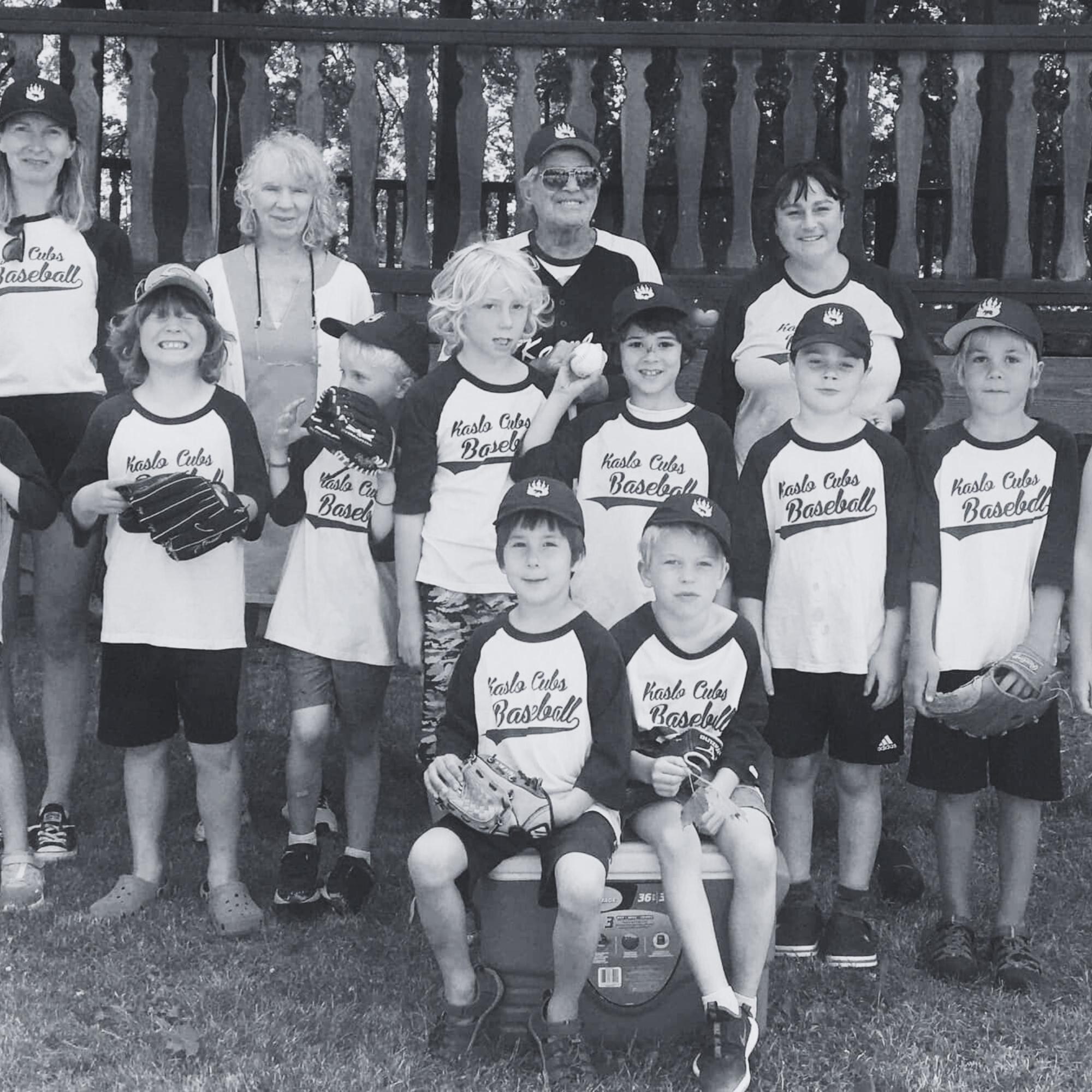A youth baseball team called 'Kaslo Cubs Baseball' with children and women, some wearing baseball uniforms, standing outdoors on grass in front of a wooden fence for a team photo.