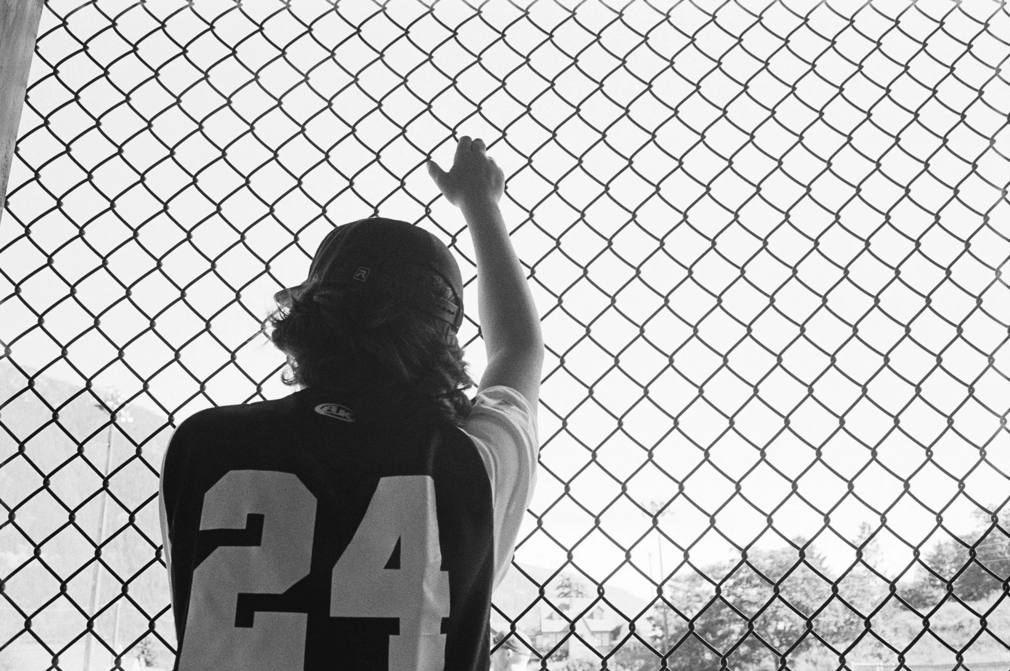Young person wearing a cap and sports jersey with the number 24, standing behind a chain-link fence, reaching up and holding onto the fence, with a background of trees and houses beyond.
