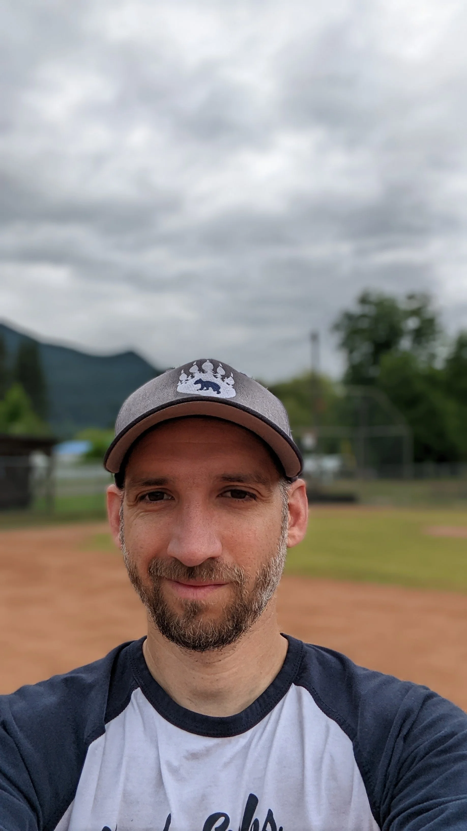 A man wearing a baseball cap and a casual t-shirt, taking a selfie outdoors on a cloudy day, with a baseball field and trees in the background.