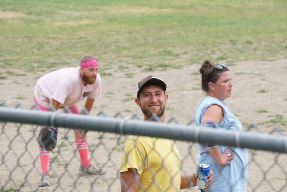 Three people at a baseball field: a man smiling and holding a can, a woman with sunglasses on her head standing with her hands on her hips, and another man crouching in the background wearing a pink outfit with knee-high pink socks and a pink headband.