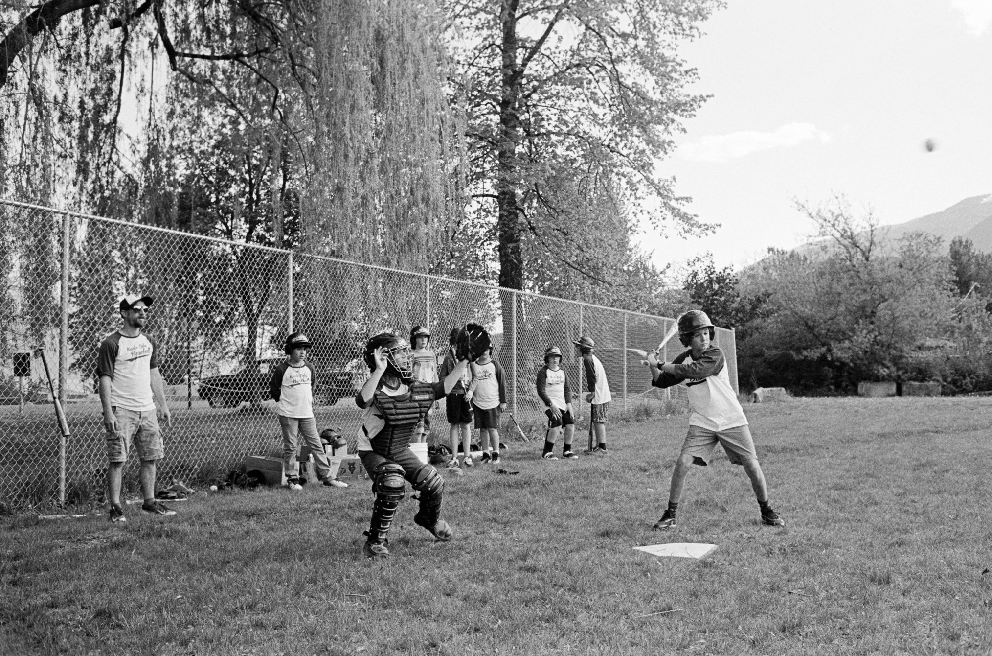 Kids playing baseball on a field with a chain-link fence, one kid batting and a girl catching with her glove, other kids and a man on the sidelines, trees and mountains in the background.