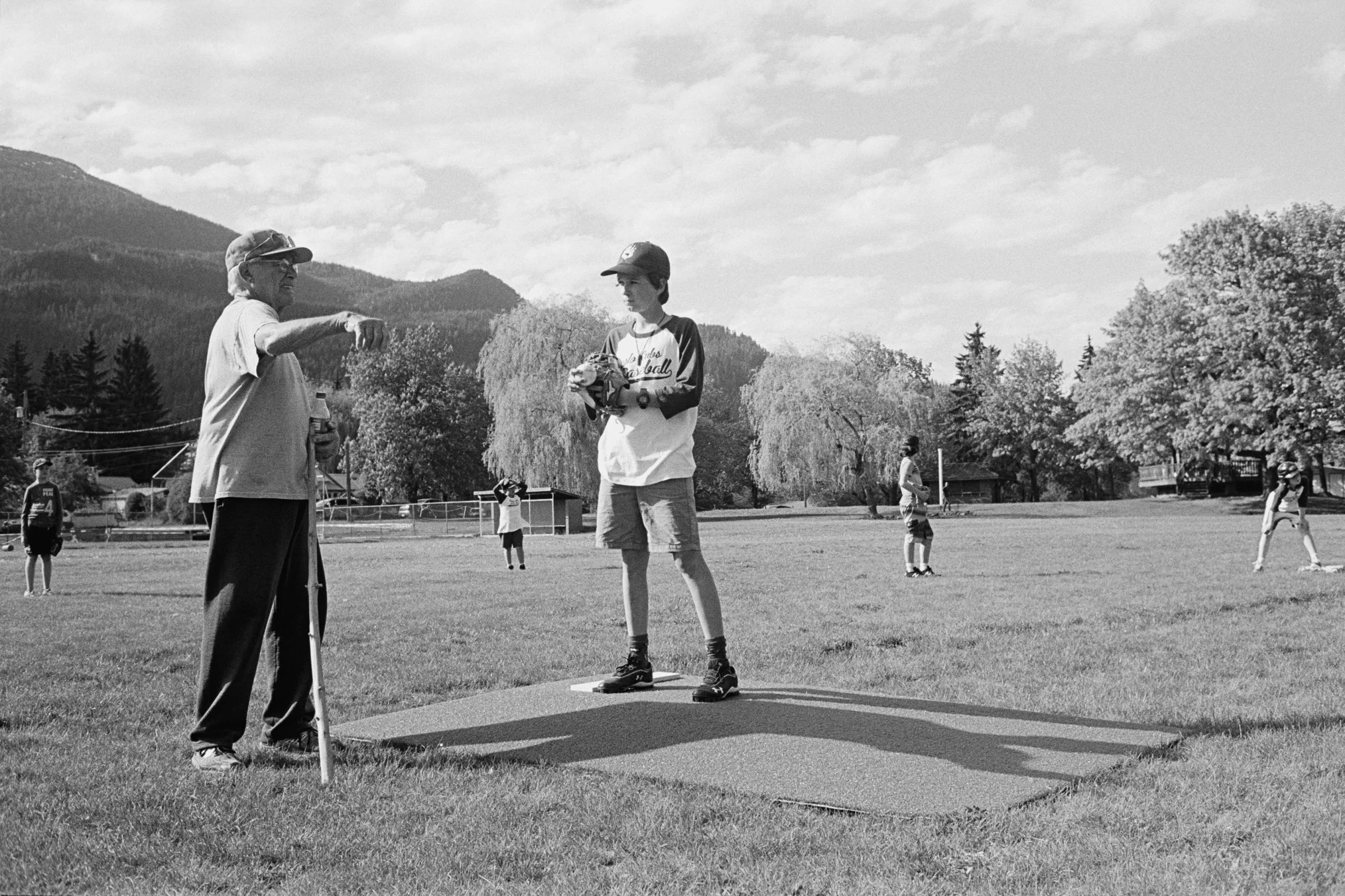 A coach talking to a young baseball player standing on a pitching mound at a baseball field, with other players and trees in the background.