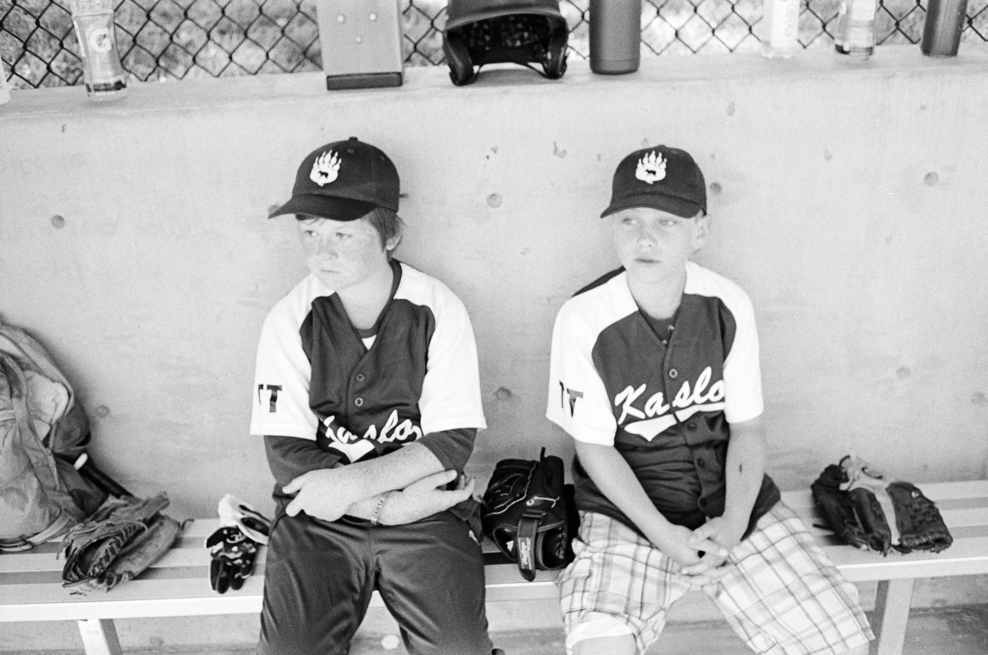 Two young boys in baseball uniforms sitting on a bench, with baseball gloves and gear around them.