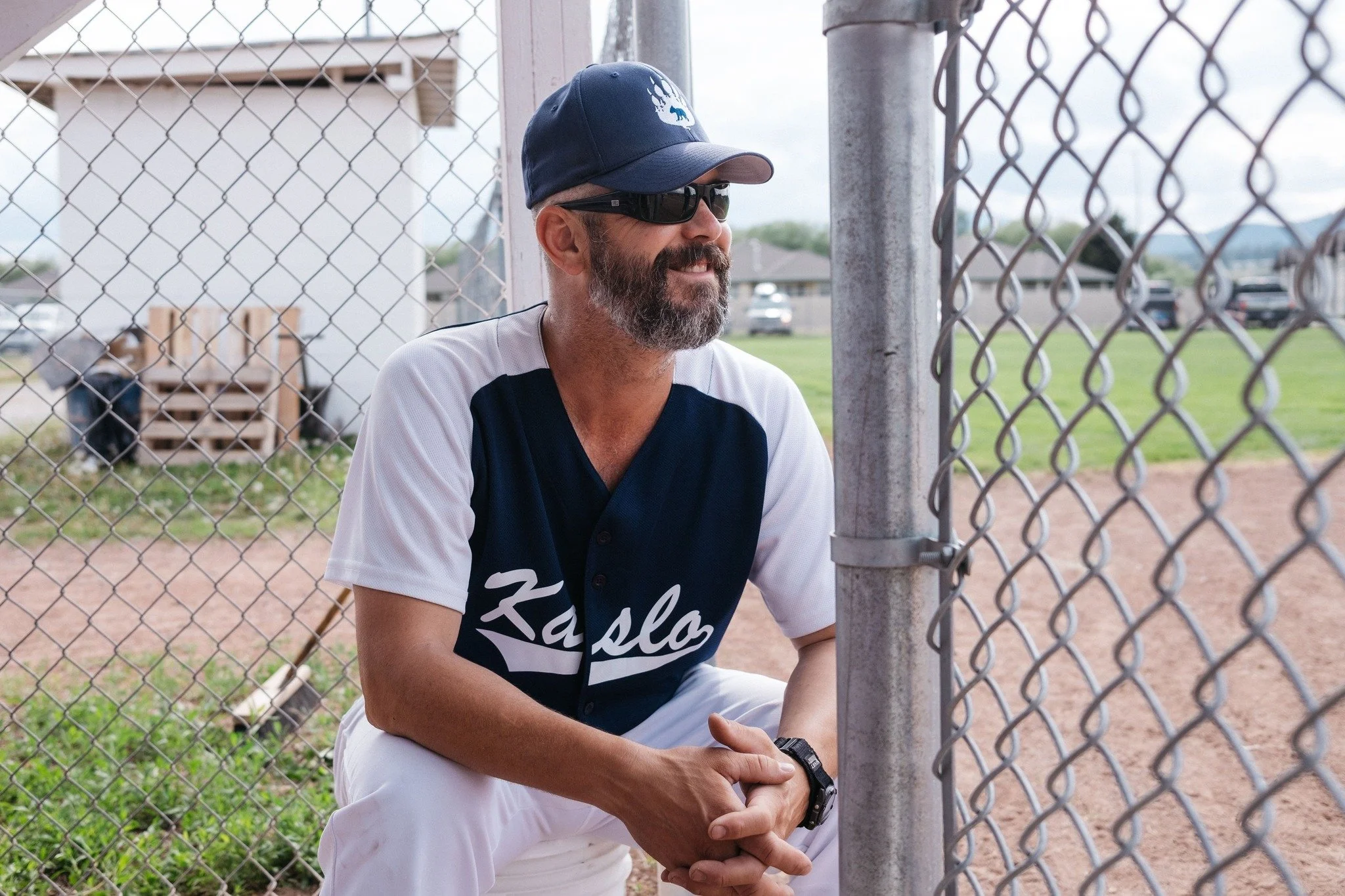 A man with a beard, wearing a navy blue cap, sunglasses, a blue and white baseball jersey, and white pants, sitting on his knees next to a chain-link fence on a baseball field.