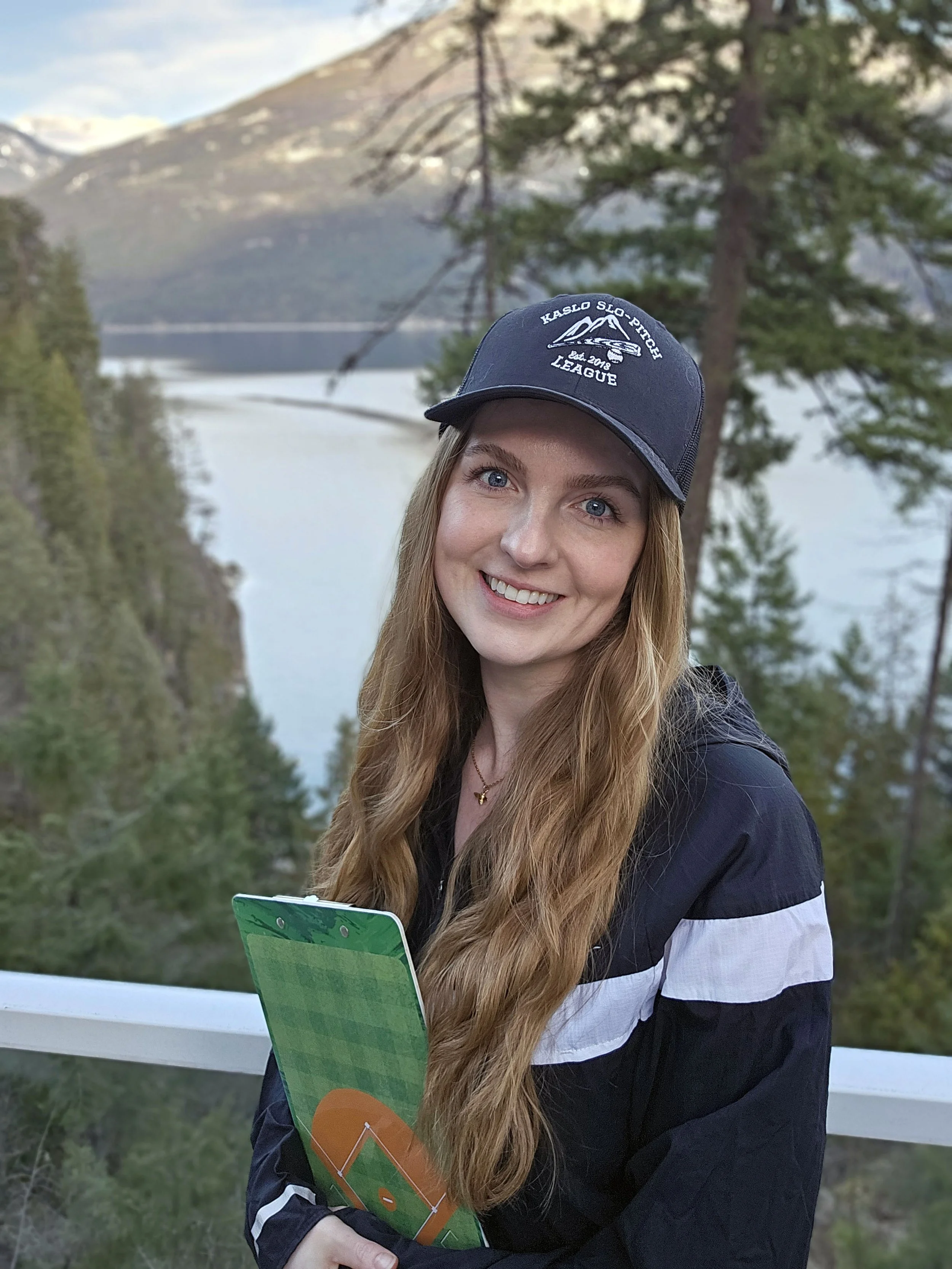 A woman with long red hair smiling outdoors near a lake and forest, wearing a navy blue cap and jacket, holding a small chalkboard with a gardening diagram.