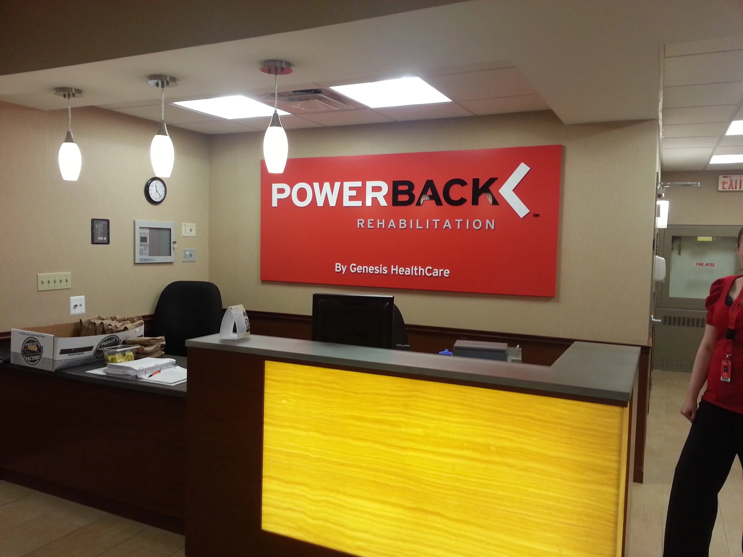 Reception area with a large red and black sign reading "PowerBack Rehabilitation by Genesis Healthcare," a desk with papers, a wall clock, and ceiling lights. A person in a red shirt is partially visible on the right.