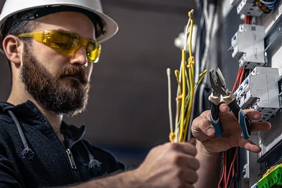 A male electrician with a beard and safety glasses working on electrical panel, using pliers to connect wires.