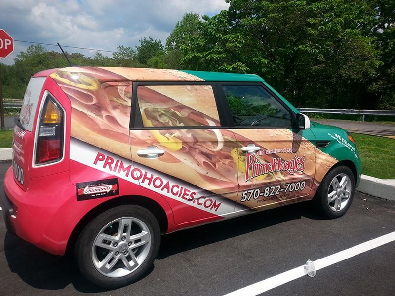 Car with a wrap advertising Primo Hoagies, featuring a large sandwich graphic on the side, the website primohogies.com, and the phone number 570-822-7000.