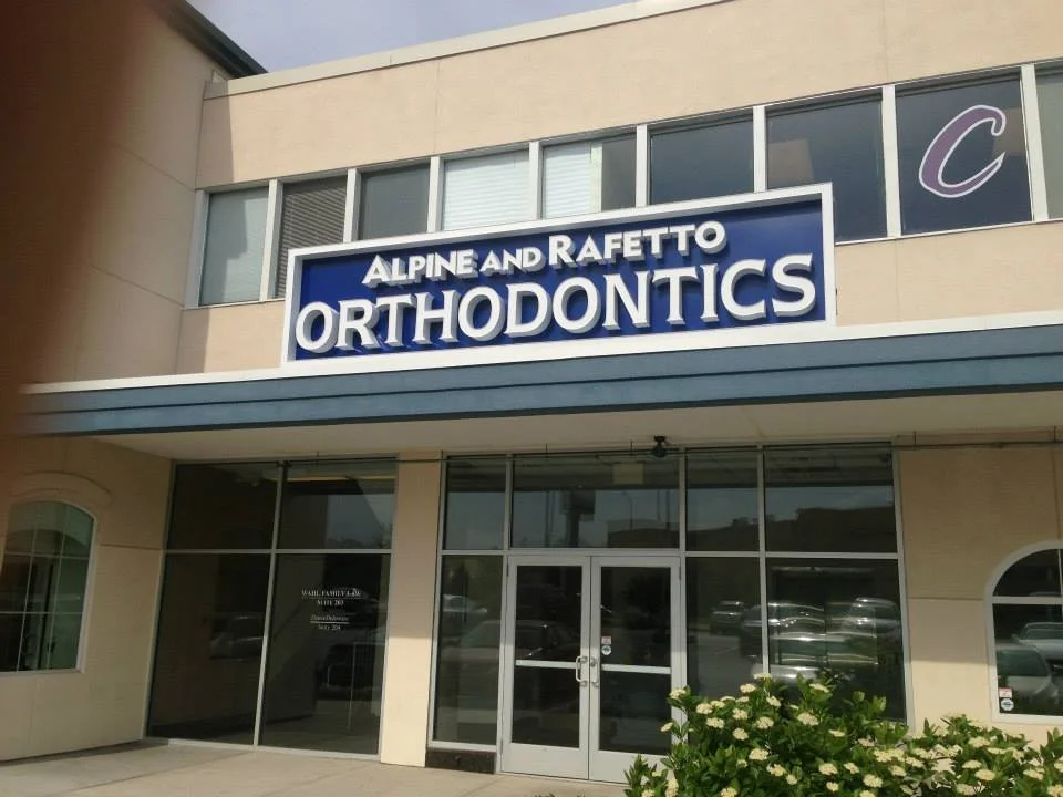 Exterior view of a building with a sign reading 'Alpine and Rafetto Orthodontics' above the entrance, with large windows and a small landscaped area with plants in front.