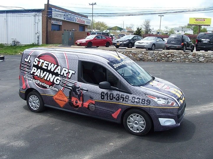 A gray commercial van with advertising for Stewart Paving parked in a lot, with several other cars and a building in the background.