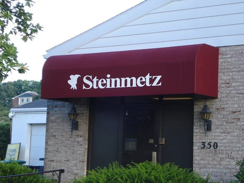 Exterior of a building with a maroon awning displaying the sign 'Steinmetz' and a dog logo, along with a black door and two wall-mounted lanterns.