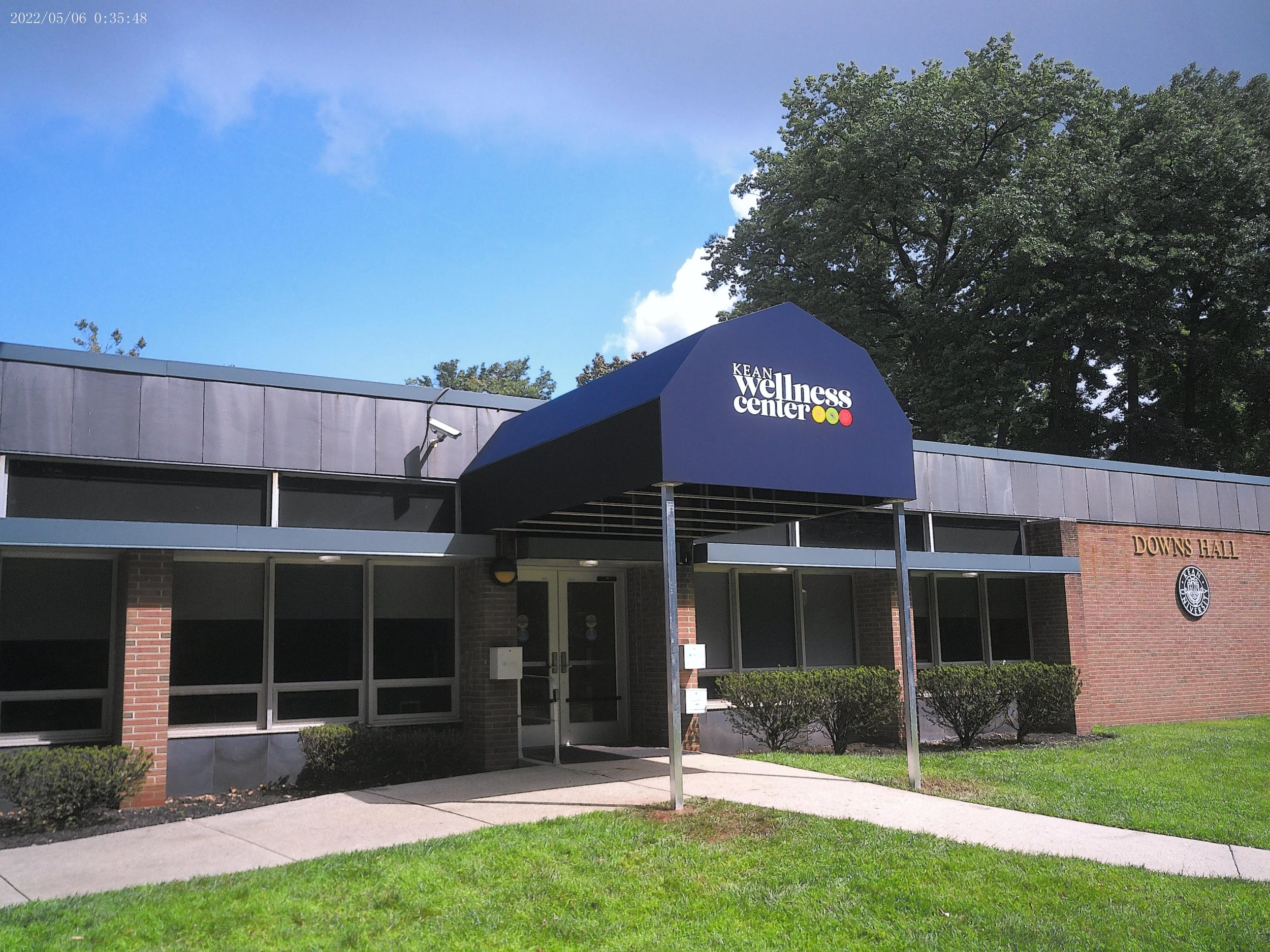 Exterior view of Kean Wellness Center building with a lush green lawn, bushes, a sidewalk, and large trees in the background. A blue sign with white text and colorful dots is visible at the entrance.