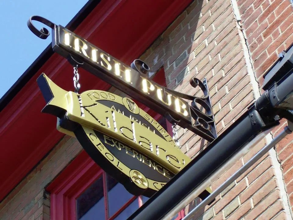 A hanging sign for a restaurant called 'Pizzeria' with a gold and black design, and a smaller sign above it that reads 'TRISTAR PUB' with illuminated letters, attached to a brick wall.