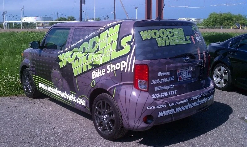 Purple van with green and white advertising decals for Wooden Wheels Bike Shop, featuring website, phone numbers, and location details, parked in a lot.