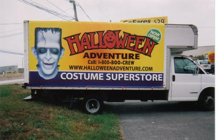 A truck advertising the Halloween Adventure costume superstore with a large cartoon of a scary clown face on its side.