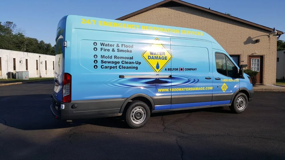 Blue service van for water damage restoration with company info and list of emergency services on it, parked on a street with buildings and trees in the background.
