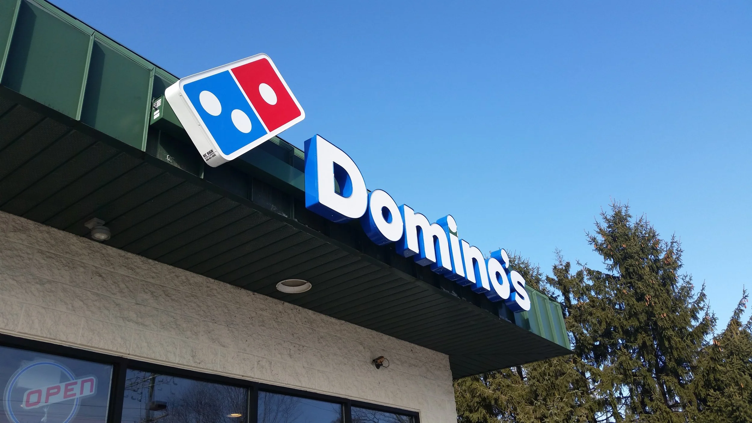 Exterior view of a Domino's Pizza store with a sign showing the Domino's logo and part of the store's name, under a clear blue sky with a large tree nearby.