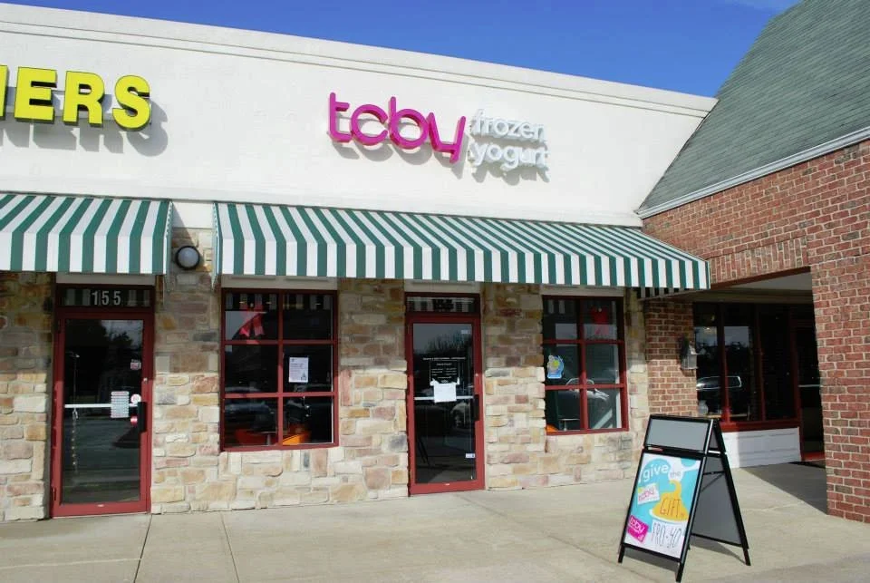 Facade of a shopping center with a Toys store, ice cream, frozen yogurt, and another shop. The storefront has a green and white striped awning, stone wall, and red window and door frames. There is a sidewalk and an easel-style sign outside.