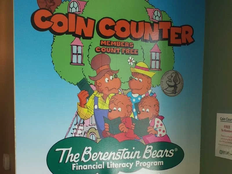 Colorful poster for the Coin Counter financial literacy program by The Berenstain Bears featuring two adults and two children reading books, with a large green tree and house in the background.