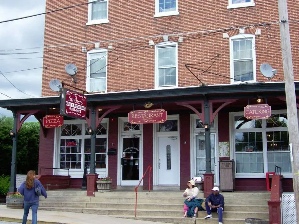 Brick building with a storefront featuring signs for a pizza restaurant and catering, and people sitting and standing outside.
